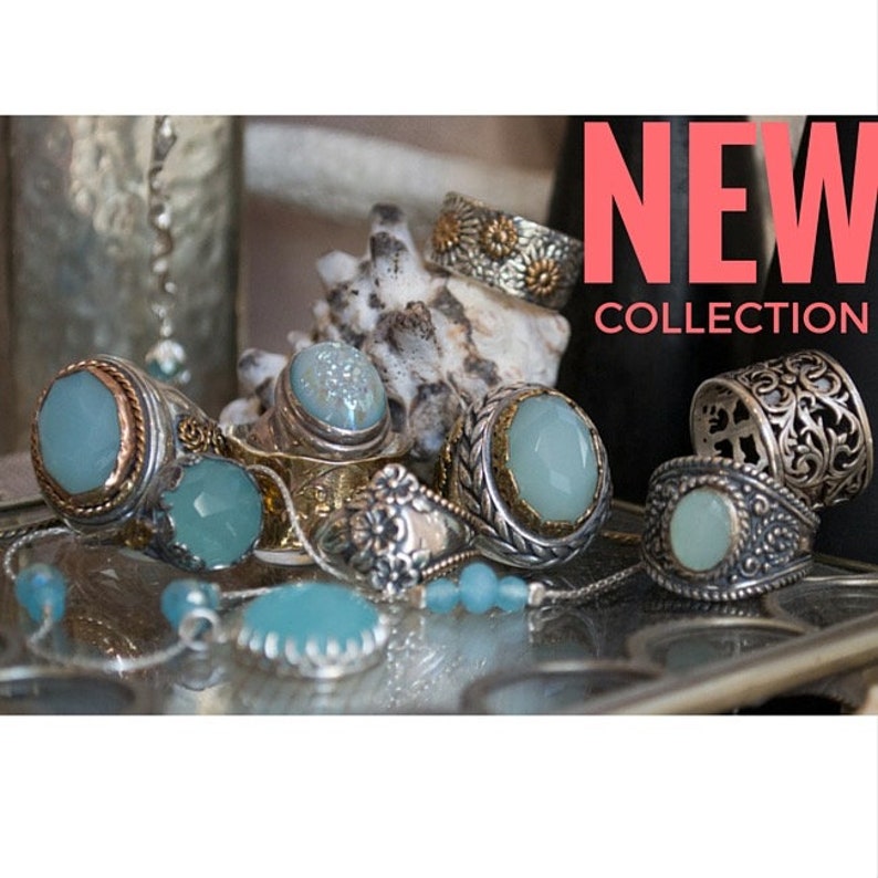 May include: A collection of silver rings with blue gemstones. The rings are arranged on a surface with a seashell. The text "NEW COLLECTION" is displayed in pink at the top of the image.