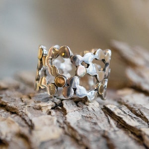 Könnte beinhalten: Ein silberner Ring mit einem filigranen, durchbrochenen Design, das ein sich wiederholendes Muster aus kleinen, abgerundeten Formen aufweist. Der Ring ist mit kleinen, goldfarbenen Punkten verziert.
