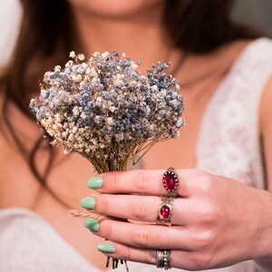 Puede incluir: Una mano con esmalte de u&ntilde;as verde sostiene un ramo de flores de aliento de beb&eacute; secas. La mano lleva dos anillos con piedras preciosas rojas.