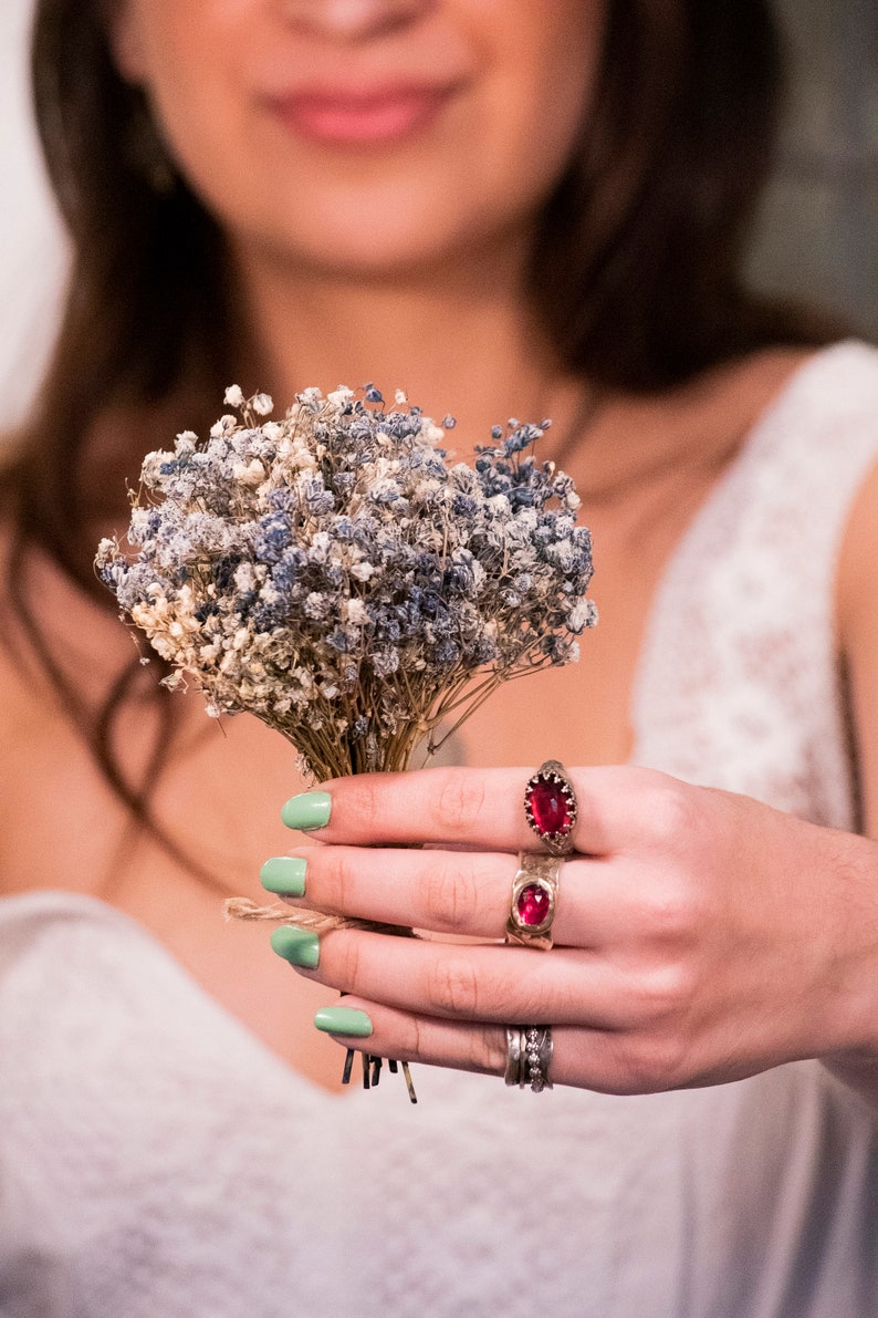 Puede incluir: Una mano con esmalte de u&ntilde;as verde sostiene un ramo de flores de aliento de beb&eacute; secas. La mano lleva dos anillos con piedras preciosas rojas.