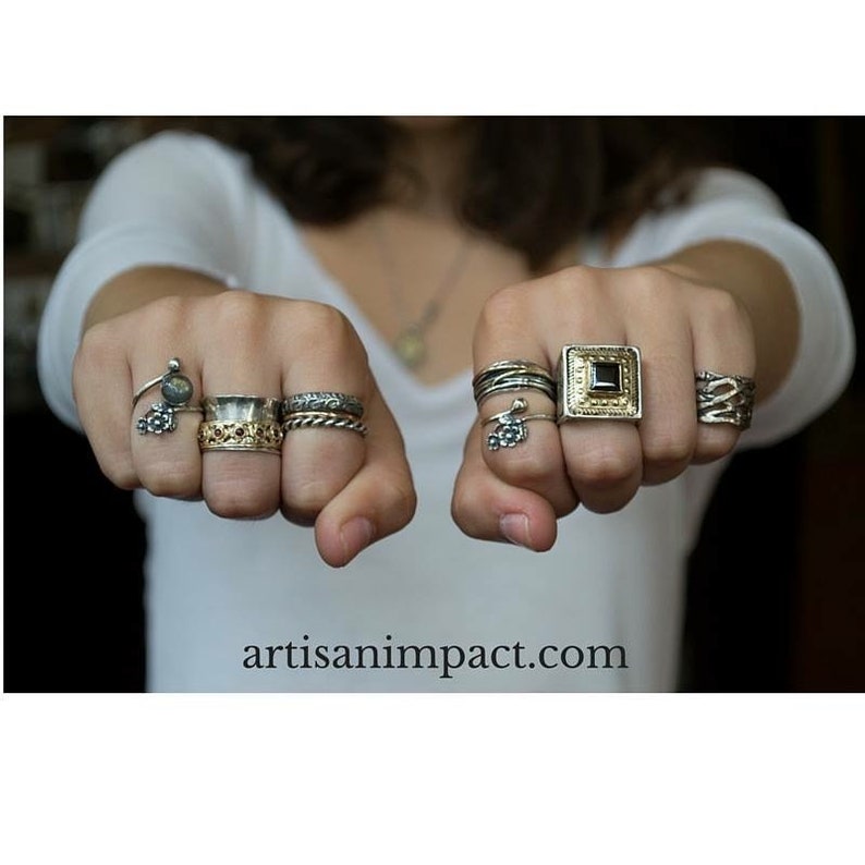 May include: A person's hands wearing a variety of silver and gold rings. The rings feature different designs, including a square ring with a black stone, a ring with a small flower charm, and a ring with a textured band.