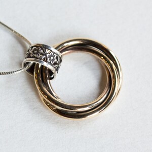 May include: A gold-toned pendant necklace featuring three interlocking rings. A silver band with a floral design encircles the rings. The necklace has a delicate silver chain. The jewelry is set against a white background.