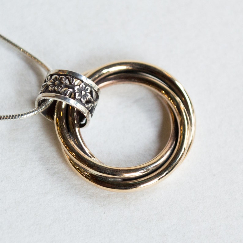 May include: A gold-toned pendant necklace featuring three interlocking rings. A silver band with a floral design encircles the rings. The necklace has a delicate silver chain. The jewelry is set against a white background.