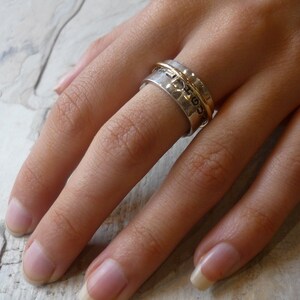 May include: A silver and gold spinning ring with a textured band. The ring is worn on a finger.