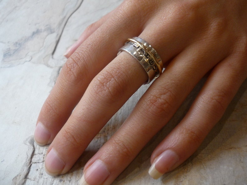 May include: A silver and gold spinning ring with a textured band. The ring is worn on a finger.
