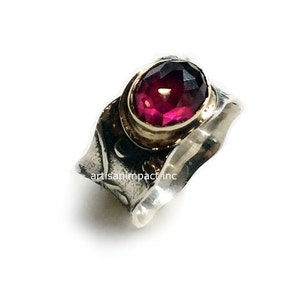 May include: A silver ring with a textured band and a deep red gemstone set in a gold bezel. The ring is handcrafted and has a unique, organic design.