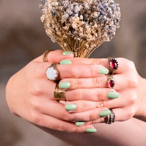 Puede incluir: Manos con esmalte de u&ntilde;as verde claro sosteniendo un ramo de flores secas blancas y moradas. Las manos llevan varios anillos de oro, incluyendo uno con una gran piedra de &oacute;palo blanco y otro con una piedra preciosa roja.