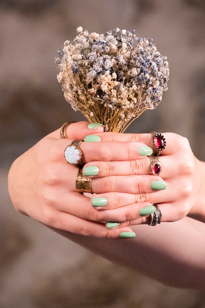 Puede incluir: Manos con esmalte de u&ntilde;as verde claro sosteniendo un ramo de flores secas blancas y moradas. Las manos llevan varios anillos de oro, incluyendo uno con una gran piedra de &oacute;palo blanco y otro con una piedra preciosa roja.