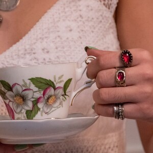 Puede incluir: Una taza de t&eacute; blanca y un platillo con un dise&ntilde;o floral. La taza de t&eacute; tiene un borde dorado y el platillo tiene un borde festoneado. La taza de t&eacute; est&aacute; siendo sostenida por una mano que lleva tres anillos con piedras preciosas rojas.