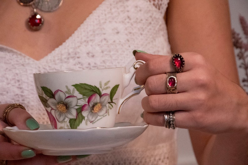 Puede incluir: Una taza de t&eacute; blanca y un platillo con un dise&ntilde;o floral. La taza de t&eacute; tiene un borde dorado y el platillo tiene un borde festoneado. La taza de t&eacute; est&aacute; siendo sostenida por una mano que lleva tres anillos con piedras preciosas rojas.