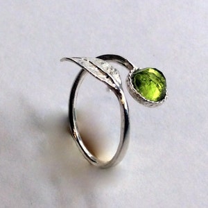 May include: A silver ring featuring a leaf motif and a round, light green gemstone. The ring's band curves, with a leaf detail and the gemstone. The ring is set against a white background.