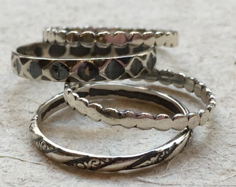 May include: Four silver rings with different designs. The rings are stacked on top of each other. The top ring has a diamond pattern, the second ring has a scalloped edge, the third ring has a scalloped edge, and the bottom ring has a floral design.