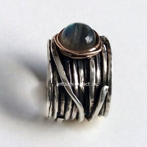 May include: A silver ring featuring a labradorite gemstone set in a rose gold bezel. The ring is crafted from multiple strands of silver wire that are twisted and woven together.
