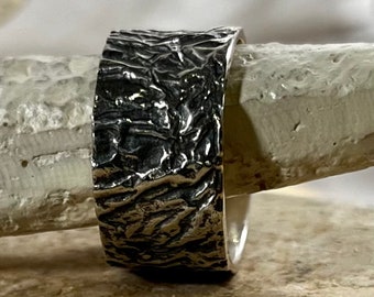 May include: A wide silver band ring with a textured, organic design. The ring has a rough, hammered finish.