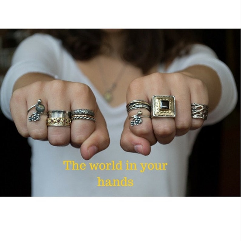 May include: A person's hands wearing a variety of silver and gold rings. The rings are stacked on each finger and feature different designs, including a square ring with a black stone, a ring with a flower design, and a ring with a textured band. The caption reads "The world in your hands."