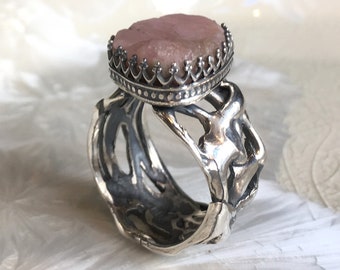 May include: A silver ring with a pink stone set in a crown-like bezel. The ring band is intricately designed with a textured, organic pattern.