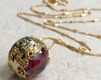 May include: A gold chain necklace with a red gemstone pendant. The pendant is surrounded by a gold filigree design.