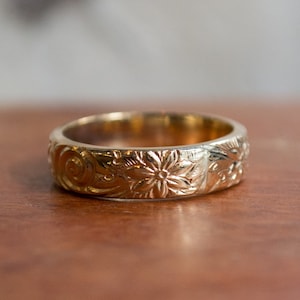 May include: A gold-colored ring with an engraved floral pattern. The ring features detailed carvings of flowers and swirling leaf designs. The ring is set on a wooden surface, showcasing its intricate craftsmanship.