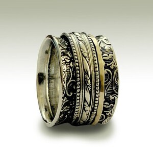 Può includere: Un anello d'argento con una fascia larga che presenta più disegni intricati. L'anello ha una fascia sottile d'oro incassata al centro.