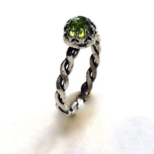 Könnte beinhalten: Ein silberner Ring mit einem gedrehten Band und einem grünen Edelstein, der in einem floralen Design gefasst ist.