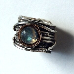 May include: A silver ring featuring a round, iridescent labradorite gemstone set in a copper bezel. The ring band is crafted from multiple strands of twisted silver wire.