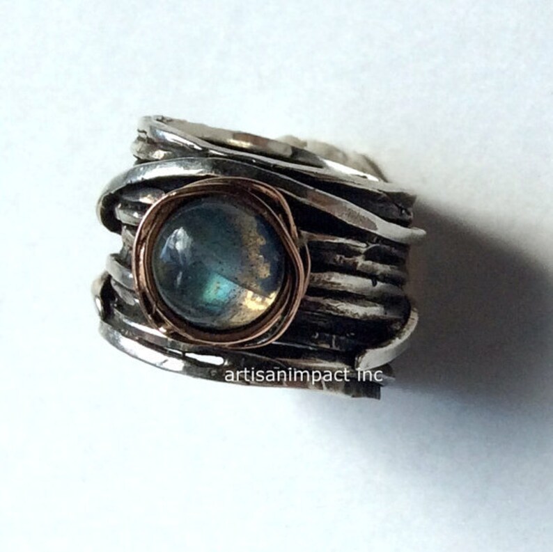May include: A silver ring featuring a round, iridescent labradorite gemstone set in a copper bezel. The ring band is crafted from multiple strands of twisted silver wire.