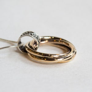 May include: A silver chain necklace with a pendant featuring three gold-colored interlocking rings and a silver ring with a decorative pattern. The pendant is a symbol of unity and connection.