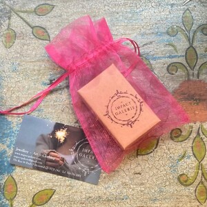 May include: A small pink organza gift bag with a brown box inside. The box has a white label with the text "IMPACT GALERIE" in a circle.