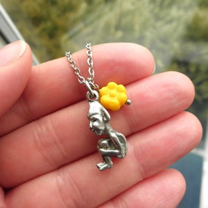 Puede incluir: Un collar de plata con una pequeña figura detallada de un gnomo sentado y un dije de flor amarilla.