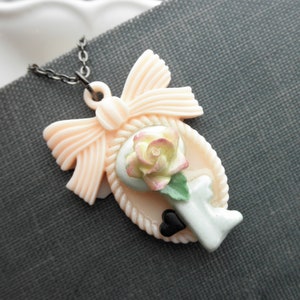 Op de afbeelding: Een roze strik met een sleutelhanger en een pastelroze en gele roos. De sleutelhanger heeft een zwart hart en een lichtblauw en groen ontwerp.