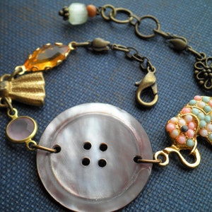 May include: A gold and brown bracelet with a large, gray, four-hole button charm, a pink and white elephant charm, and a variety of other charms, including a bow, a clear bead, and an orange bead.