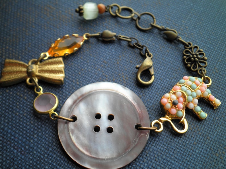 May include: A gold and brown bracelet with a large, gray, four-hole button charm, a pink and white elephant charm, and a variety of other charms, including a bow, a clear bead, and an orange bead.