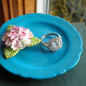 May include: A turquoise blue ceramic ring dish with a white and pink porcelain flower and a silver ring with a large clear stone.