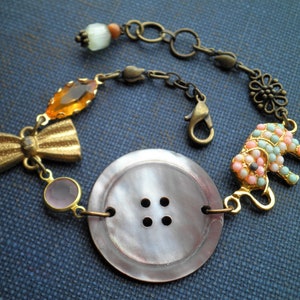 May include: A bracelet with a large, pearly button, a gold bow, a pink and orange glass bead, and a beaded elephant charm. The bracelet is made with a chain and a lobster clasp.