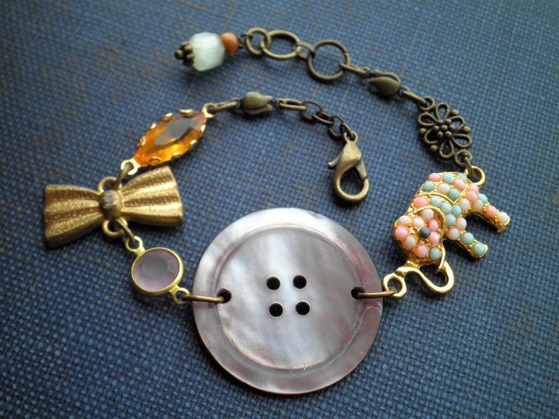 May include: A bracelet with a large, pearly button, a gold bow, a pink and orange glass bead, and a beaded elephant charm. The bracelet is made with a chain and a lobster clasp.