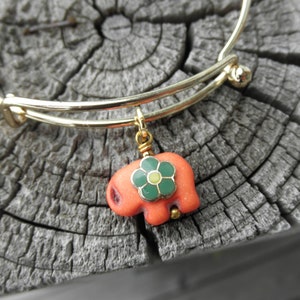 May include: A gold expandable bangle bracelet with a charm of a coral-colored elephant with a green and yellow flower on its side.