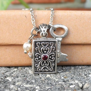 May include: A silver necklace with a small, ornate box pendant featuring a red gemstone. The pendant is attached to a key charm and a pearl charm.