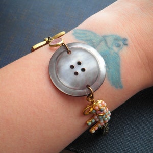 May include: A bracelet with a large, gray button charm, a pink gemstone, and a small, beaded elephant charm. The bracelet is on a person's wrist.