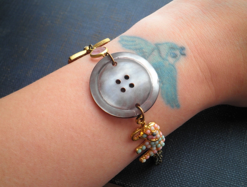 May include: A bracelet with a large, gray button charm, a pink gemstone, and a small, beaded elephant charm. The bracelet is on a person's wrist.