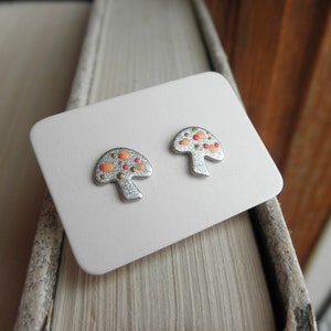 May include: A pair of silver mushroom-shaped earrings with red and gold accents. The earrings are displayed on a white card.