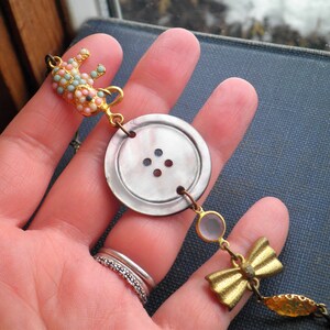 May include: A gold necklace with a large, white button charm, a smaller pink and blue elephant charm, a clear glass bead, and a gold bow charm.