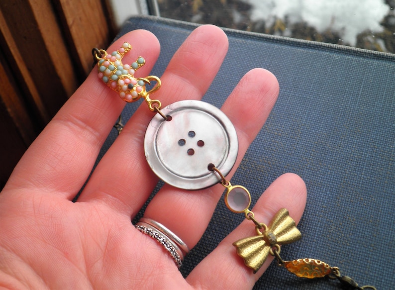 May include: A gold necklace with a large, white button charm, a smaller pink and blue elephant charm, a clear glass bead, and a gold bow charm.