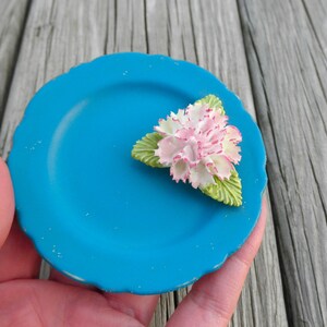 May include: A small, teal blue ceramic plate with a white and pink flower decoration. The flower has green leaves and is centered on the plate.