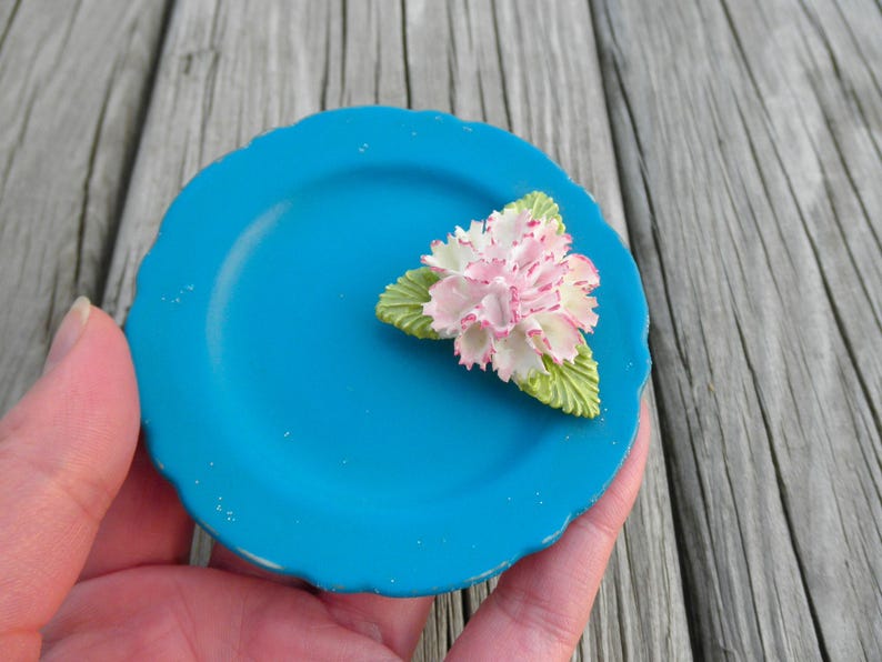 May include: A small, teal blue ceramic plate with a white and pink flower decoration. The flower has green leaves and is centered on the plate.