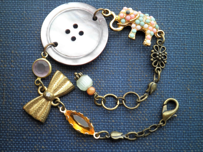 May include: A charm bracelet with a large, brown button, a gold bow, a pastel pink and blue elephant charm, and a variety of other beads and charms.