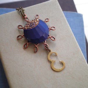 May include: A handcrafted necklace featuring a faceted, deep blue pendant with copper wire detailing. The pendant is connected to a chain and a gold-colored letter "E" charm. The necklace rests on a neutral-toned surface.