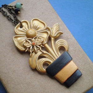 May include: Gold-tone necklace featuring a flower with a bee charm and a black and gold striped pot pendant.