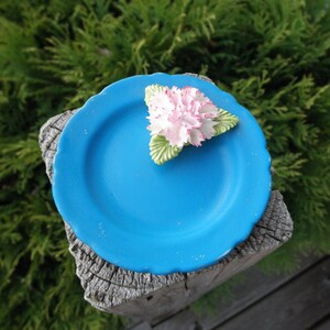 May include: A small blue ceramic plate with a white flower decoration. The plate is sitting on a wooden surface.