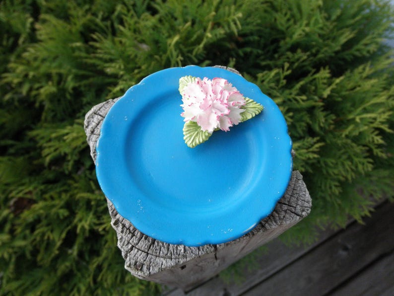 May include: A small blue ceramic plate with a white flower decoration. The plate is sitting on a wooden surface.