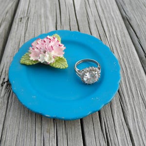May include: A blue ceramic ring dish with a white and pink flower and a silver ring with a large clear stone.
